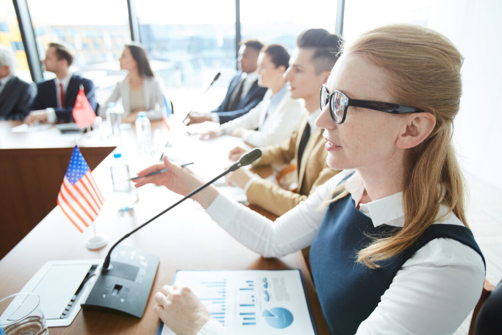 Municipal official speaking into a microphone during a formal council meeting with charts and documents on the table illustrating CAP scaling for municipalities in a multi-stakeholder governance setting.