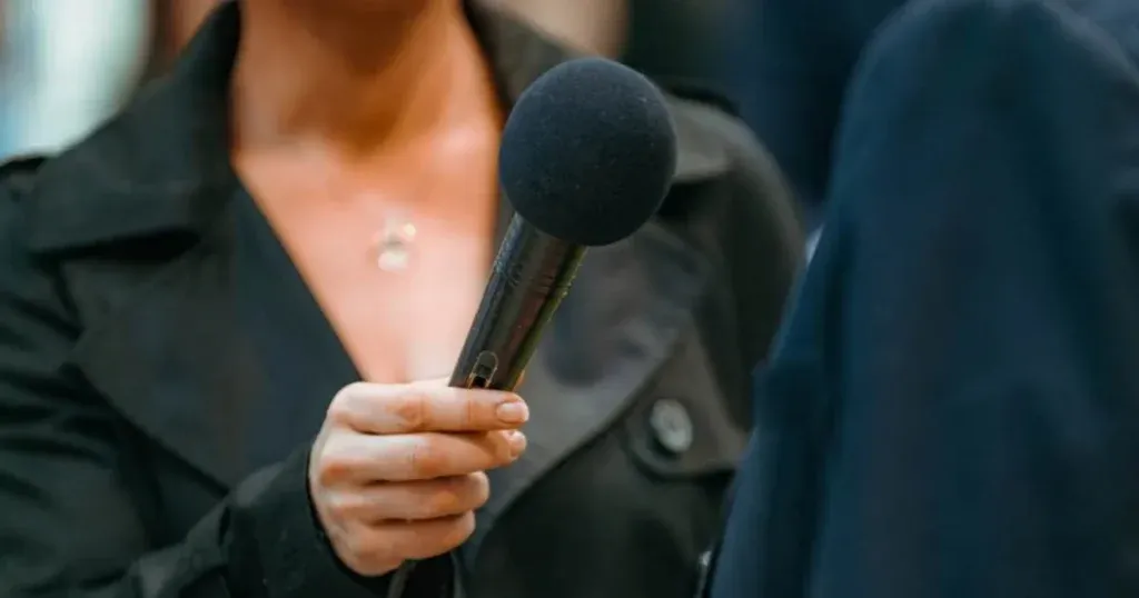 Reporter holding a black handheld microphone toward an interview subject, focus on the microphone and hand.