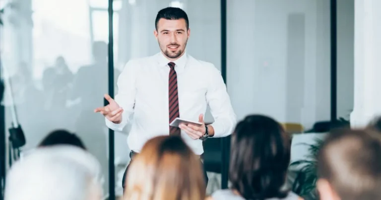 Business professional presenting to a seated audience in a modern office conference room.