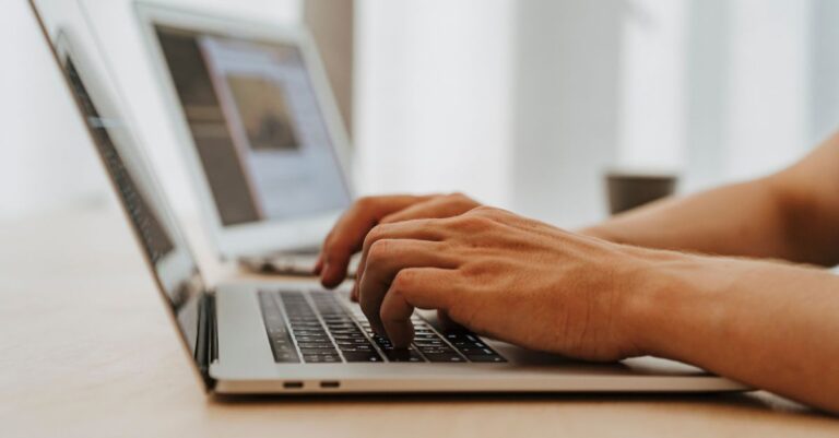 Close-up of a person typing on a laptop with another laptop displaying a webpage in the background.