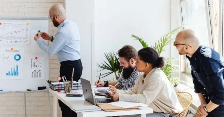 Four professionals collaborate during a business meeting, reviewing financial charts and graphs displayed on a presentation board in a bright office. Is this conversation helpful so far?