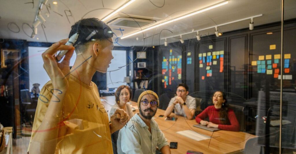 Startup team collaborating around a conference table while one person sketches ideas on a glass wall covered with notes and diagrams.