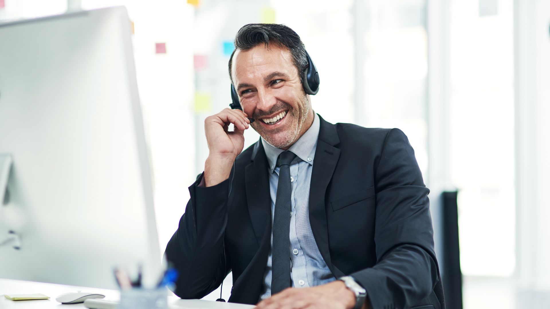 A smiling man in a suit wearing a headset, sitting at a desk and speaking during a enterprise sales call in a bright, modern office setting.
